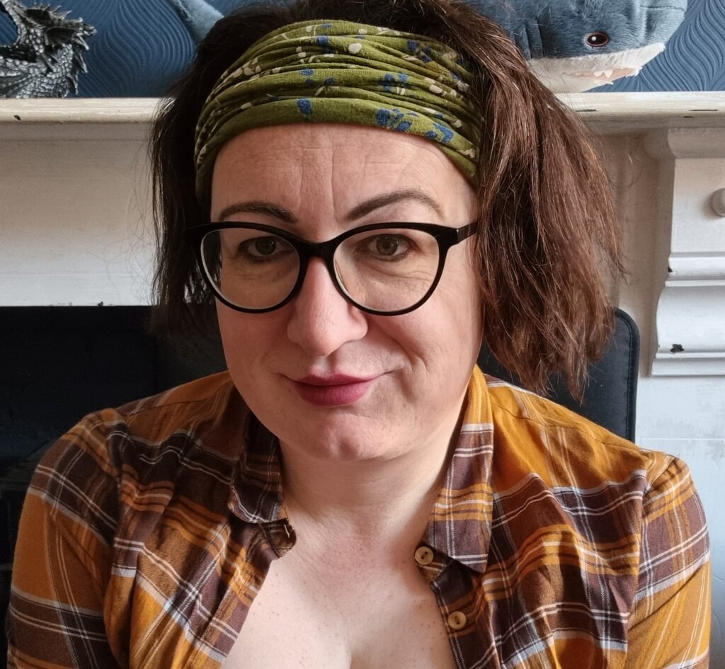 Photo of person-centred therapist, Laurie Atkinson. They are facing off to the side of the camera and are wearing black-framed glasses, a checked yellow shirt and a green hairband. In the background, an IKEA Blåhaj shark is just visible.